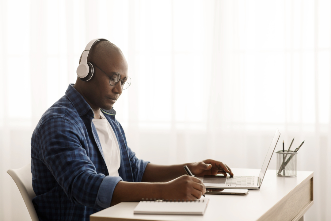 man wearing headphones writing notes and using computer, sitting at desk near window